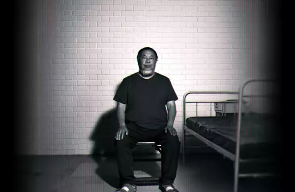 Black-and-white image of Ai Weiwei sitting on a chair beside a simple metal bed against a brick wall.