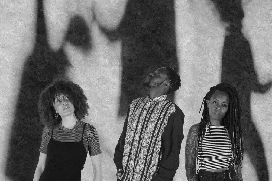 A black and white photograph of three members of The Untold Orchestra with shadowy figures in the background