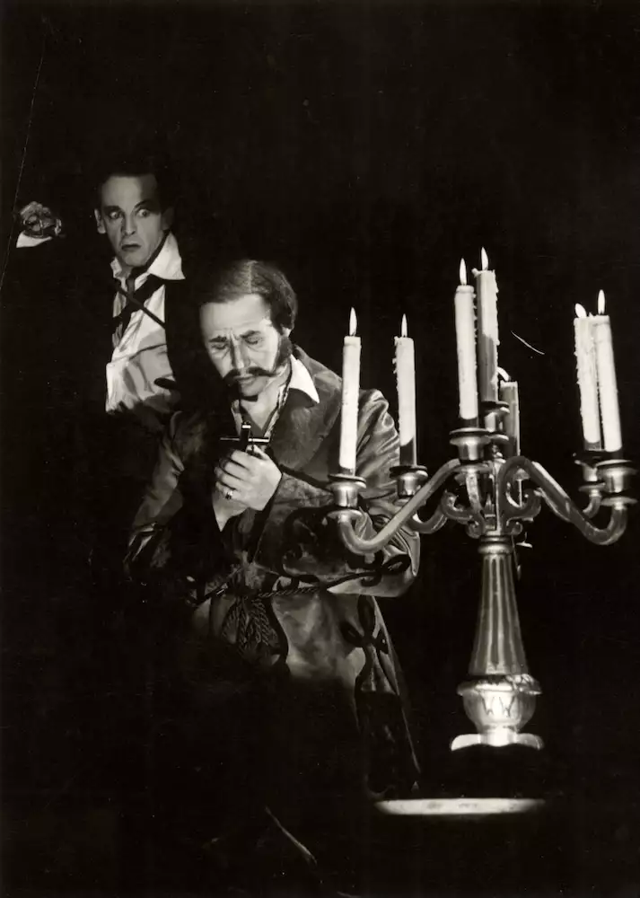 Robert Helpmann (Hamlet) sneaks up behind Anthony Quayle (Claudius), who is praying with his eyes closed, holding a cross stood next to a candle-stick. The photo is black and white.