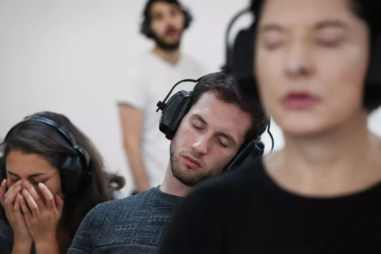 Marina Abramović with her eyes closed and headphones on, sat next to two other people doing the same.