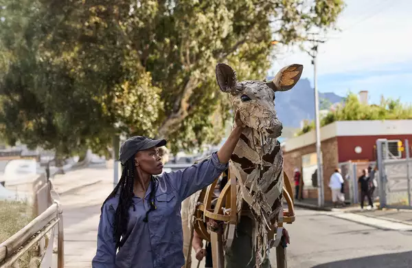 A woman in a cap touches the head of a life-sized giraffe puppet made from cardboard and wood, as part of The Herds. The background shows an urban environment with people watching and a scenic view of trees and mountains.