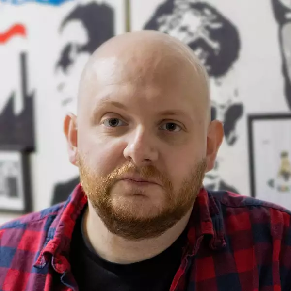 Headshot of Patrick Nicholls wearing a black t-shirt under a red and dark blue chequered open shirt.