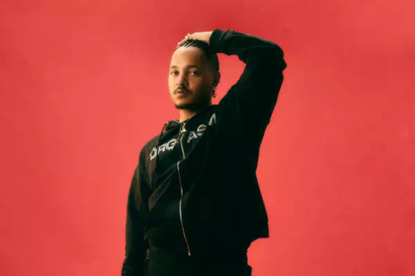 Bennettiscoming wearing a black tracksuit top, on a bright red background. They are holding a hand above their head.