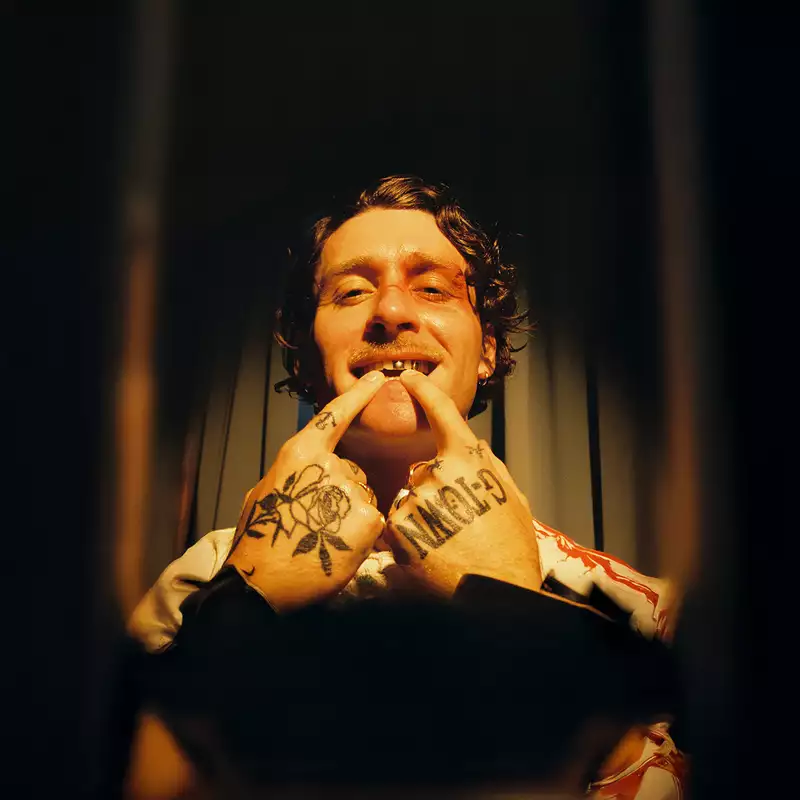Photo of Kettama on a dark brown, shadowed background. His hands are tattooed and he is pointing at his metal teeth.