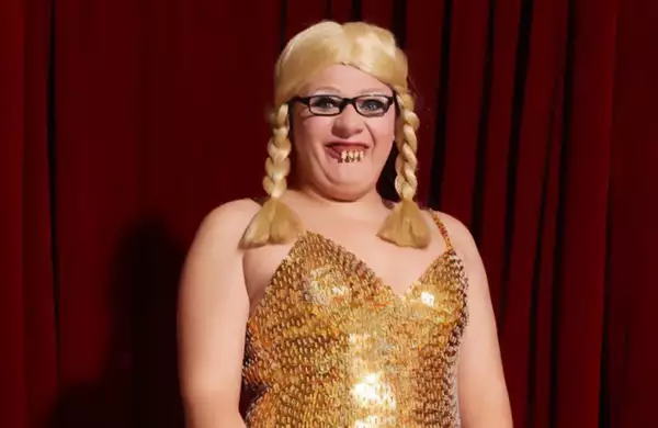 Bailey J Mills in a gold glittery dress wearing a plaited blonde wig, stood infront of red curtains