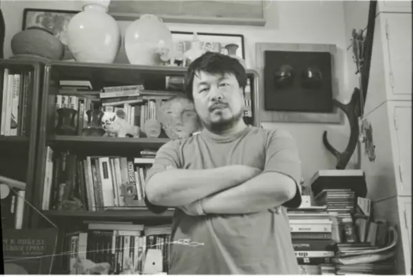 Black and white photograph of Ai Weiwei stood in front of a bookshelf