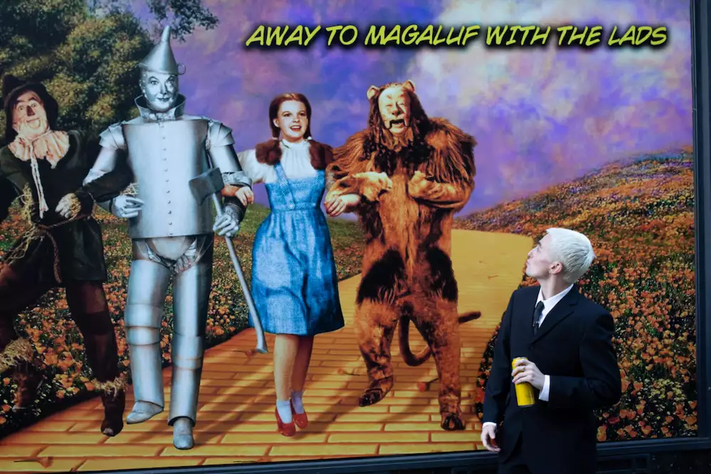 Trackie McLeod stood infront of a billboard that displays a scene from Wizard of Oz with the words 'AWAY TO MAGALUF WITH THE LADS'