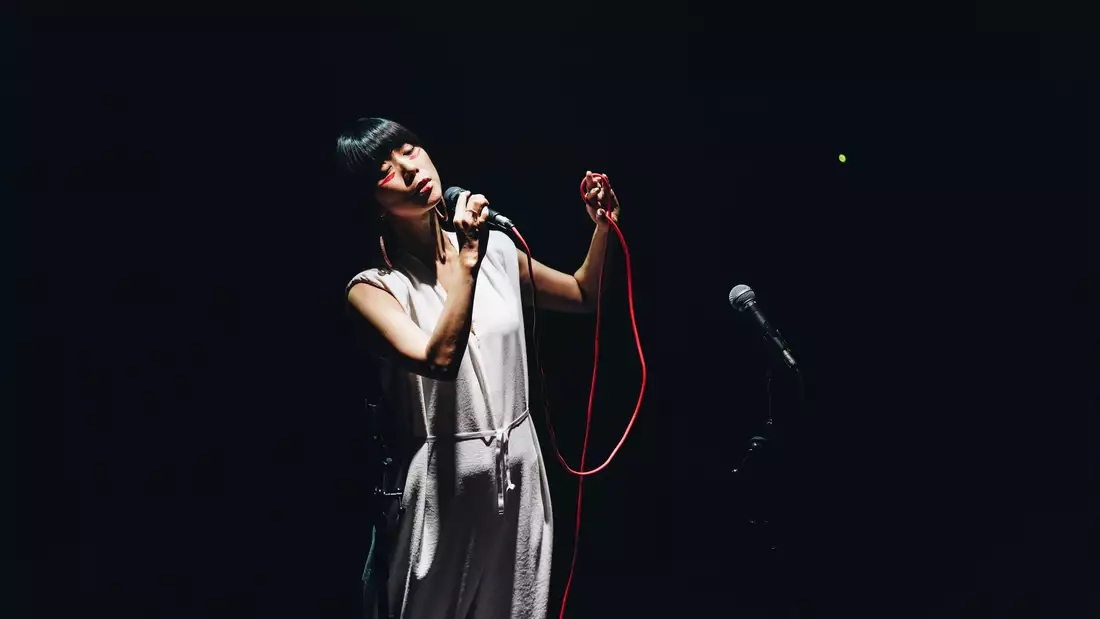 A photograph of musician Hatis Noit, standing on stage against a black background, holding a microphone with a red cable