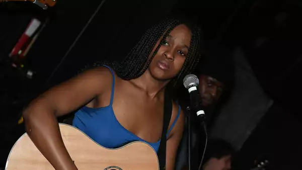 Naomi Kalu playing acoustic guitar and singing into a microphone. They are wearing a blue top.