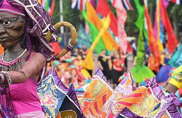 Colourful parade with large puppets, vibrant costumes and waving flags in a crowd.