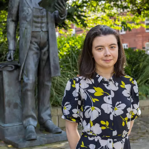 Headshot of Amelia Hall wearing a black dress with yellow and white flowers on it. They are stood outside in front of a statue and a tree.