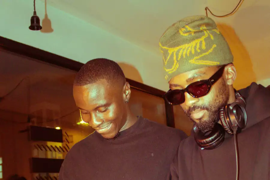 Two people stood behind decks smiling. One wears a beige and yellow hat with black sunglasses and big headphones.