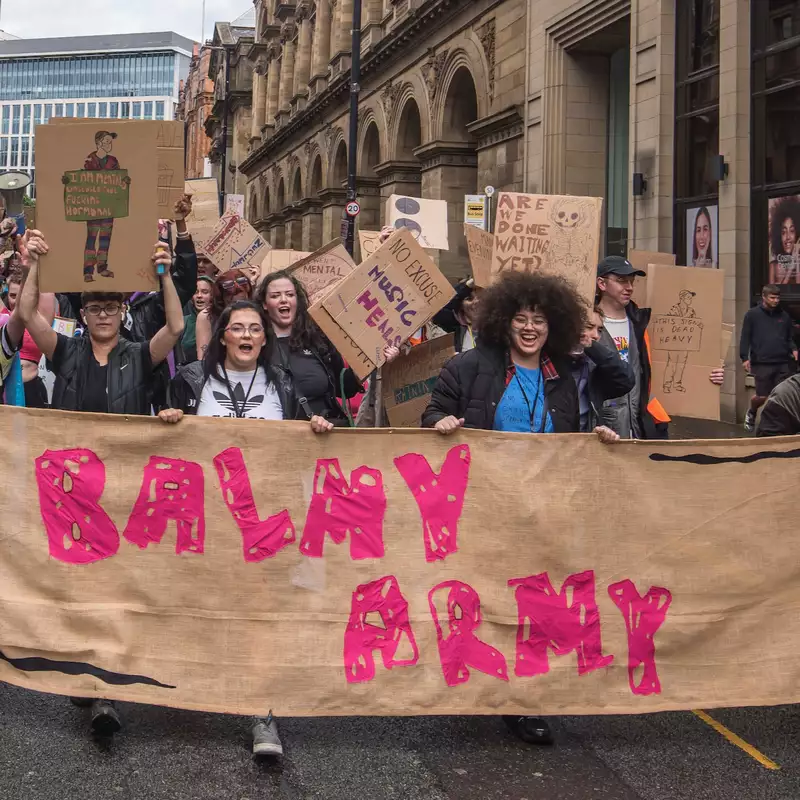 A group of young people marching through Manchester City Centre with a huge banner that reads 'Balmy Army'. Many are holding placards and megaphones.