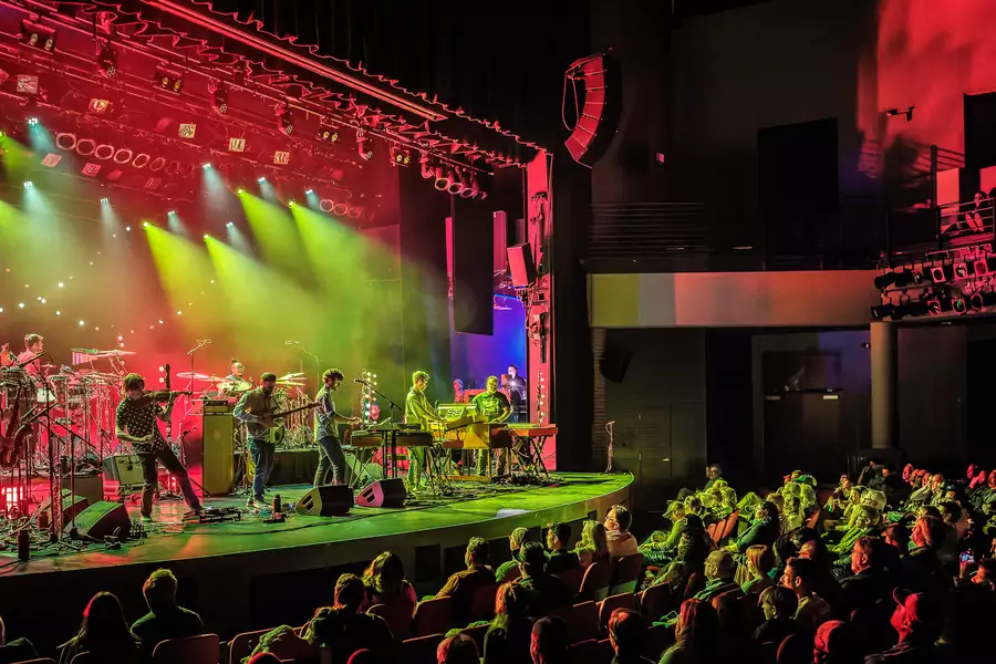The band Snarky Puppy performing on a stage to the left. Audience members are sat down watching to the right. Red and green lights shine from the stage and light up the audience sat down on the right.