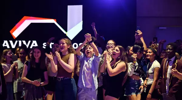 A cheering crowd of young people celebrate enthusiastically in the Social, with a glowing "Aviva Studios" sign in the background.