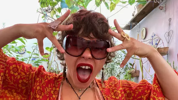 Chia wearing large sunglasses, red lipstick, and fake vampire fangs poses with fingers spread by their face. They have short curly hair, bold rings, and are dressed in a red and orange patterned top, surrounded by plants.