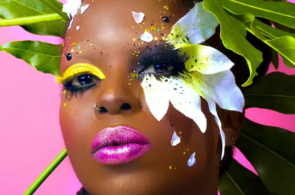 A close-up of a woman with vibrant, flower and. plant-inspired make-up. She wears bright pink lipstick. Her right eye has bright yellow eyeshadow; her left eye has black eyeshadow with petals attached to her face.