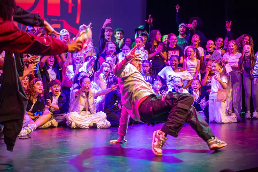 A breakdancer performs a dynamic move on stage while a large, energetic crowd of young people surrounds him, cheering, pointing, and celebrating the performance under colorful stage lights.