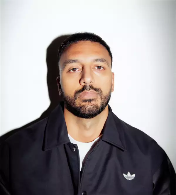 Ayman Chaudhry wearing a white t-shirt and zipped up black adidas jacket, on a white background