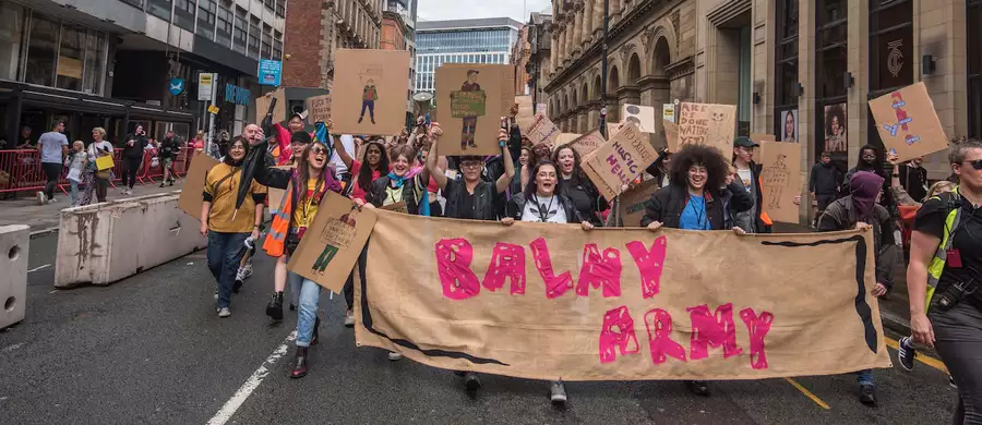 A group of young people marching through Manchester City Centre with a huge banner that reads 'Balmy Army'. Many are holding placards and megaphones.