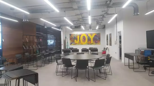 Photo of the Lab at Aviva Studios, showing an empty room with chairs, round tables and a colourful painting featuring the word 'JOY' on the wall