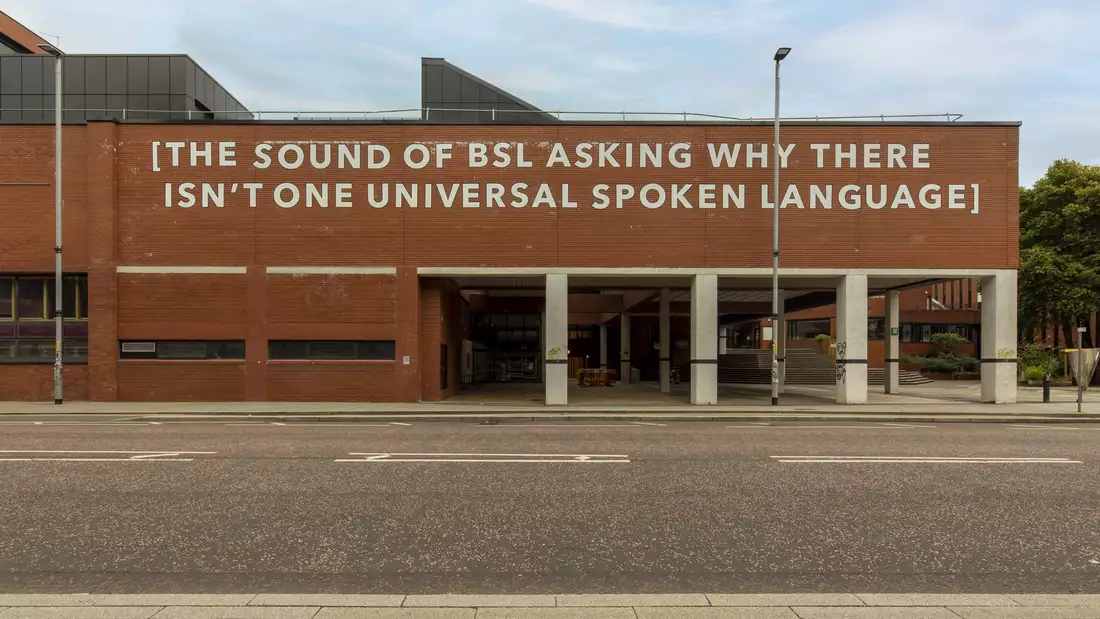 A photograph of a building with a large text installation applied with white adhesive vinyl. The text reads "[THE SOUND OF BSL ASKING WHY THERE ISN'T ONE UNIVERSAL SPOKEN LANGUAGE".