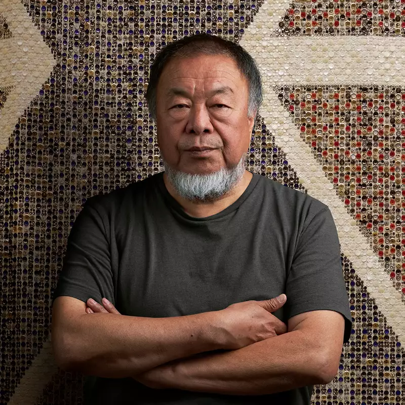 Ai Weiwei stood with arms folded wearing a black t-shirt, in front of a giant flag made of buttons