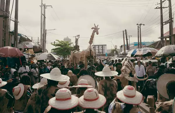 A large crowd of people at an outdoor market wearing hats. They are looking at large giraffe and elephant puppets.