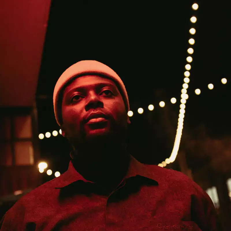 Marco Wolf wearing a shirt and beanie. It is night time and they are looking upwards. There are lights in the background.