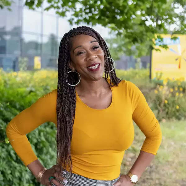 Headshot of Sharlene Small smiling and wearing a bright orange/yellow top. They are stood outside under a tree.