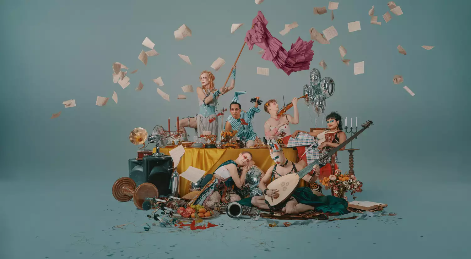 A group of performers around a table littered with objects, some are playing musical instruments, one is waving a flag