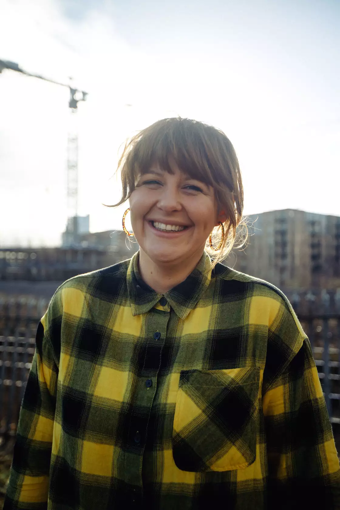 Test Card Girl photographed standing outside with flats and cranes in the background by Marieke Macklon