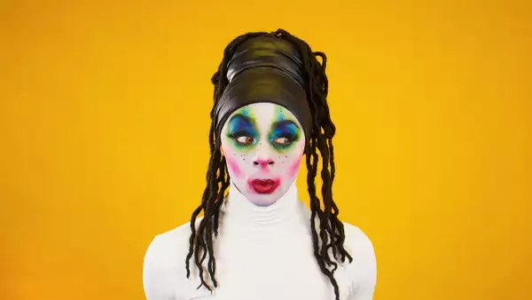 A person looking to the side wearing bright makeup and a white turtleneck top – on a bright orange background