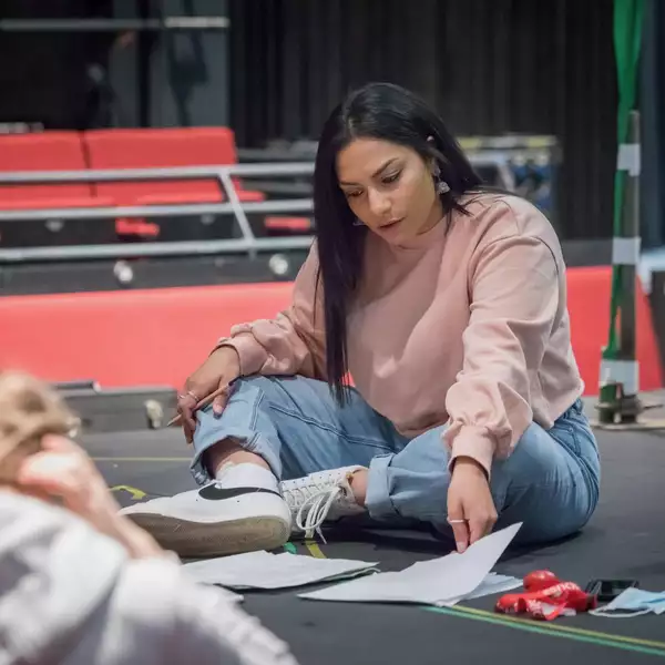 Gitika Buttoo sat on the stage floor of a theatre reading a script. She is wearing blue jeans, trainers and a light pink jumper.