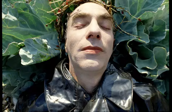 Still from Derek Jarman's The Garden (1990), showing a close up of Jarman's face. He is lying on a cabbage plant, wearing a black leather jacket and golden crown.