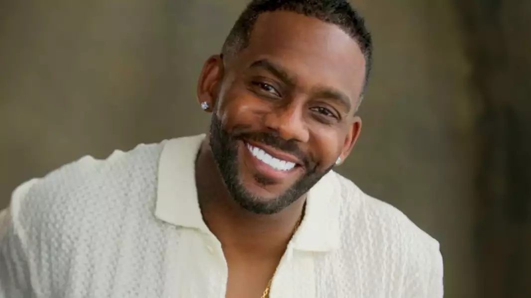 Richard Blackwood wearing an off-white textured shirt. He has short hair, a well-groomed beard and is wearing stud earrings. The background is a blurred grey/brown.