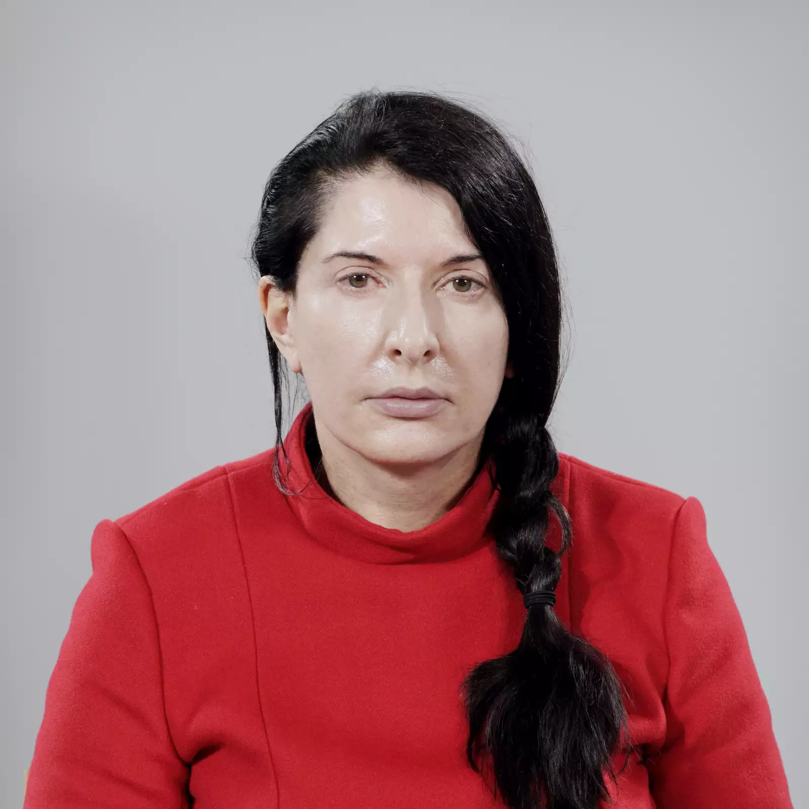 Marina Abramović looking directly into the camera during The Artist is Present. She is wearing a bright red dress.