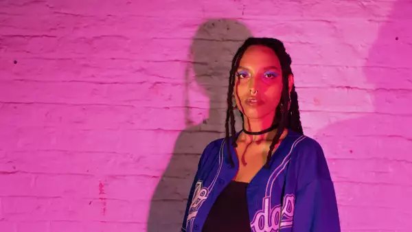 Ayy Den stood in front of a pink brick wall. Their shadow is visible on the wall behind them.