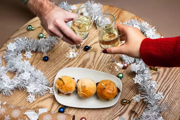 Two hands clinking champagne glasses over a festive table with small burgers on a plate, silver tinsel, and Christmas decorations.