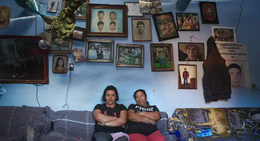 Two women sat on a sofa against a blue wall filled with photographs