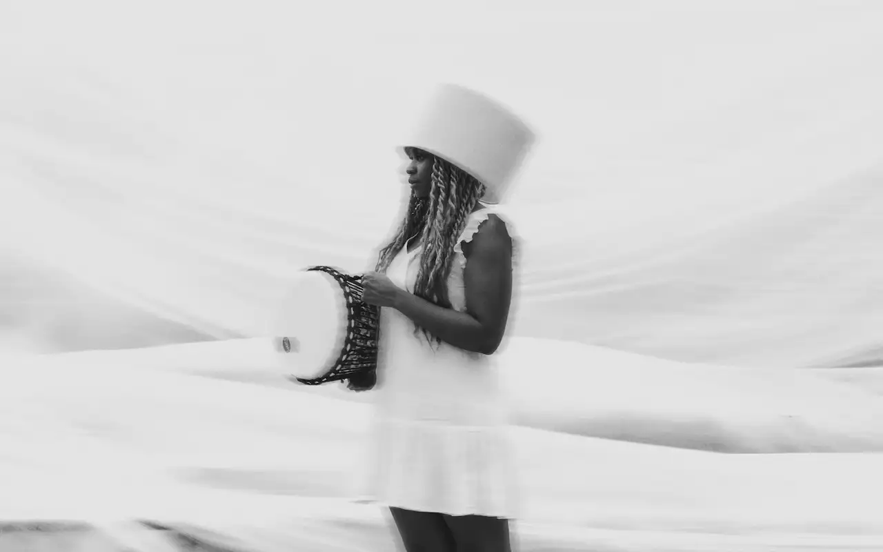 A person in a white dress and headdress carries a drum. The background is white and out of focus.