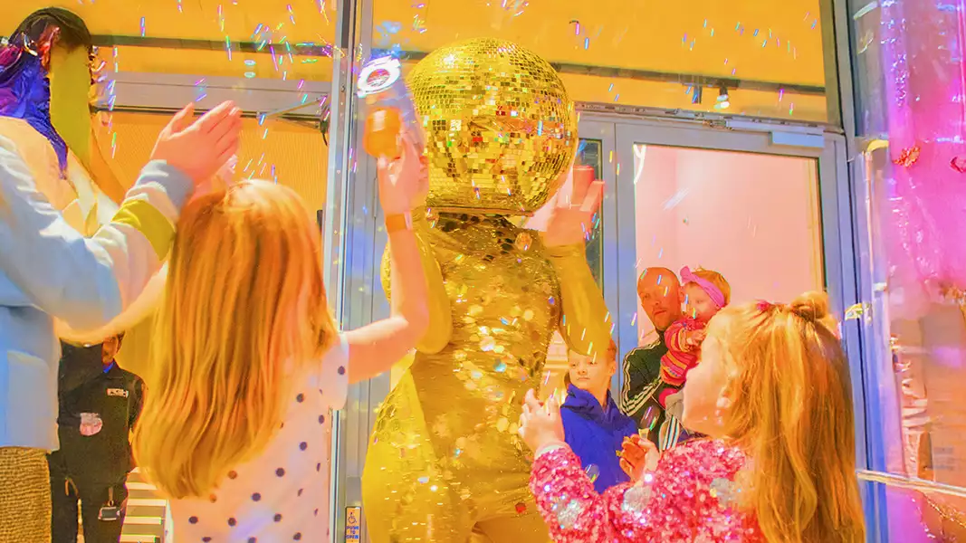A person dressed as a golden disco ball in the Social surrounded by excited children playing with bubbles.
