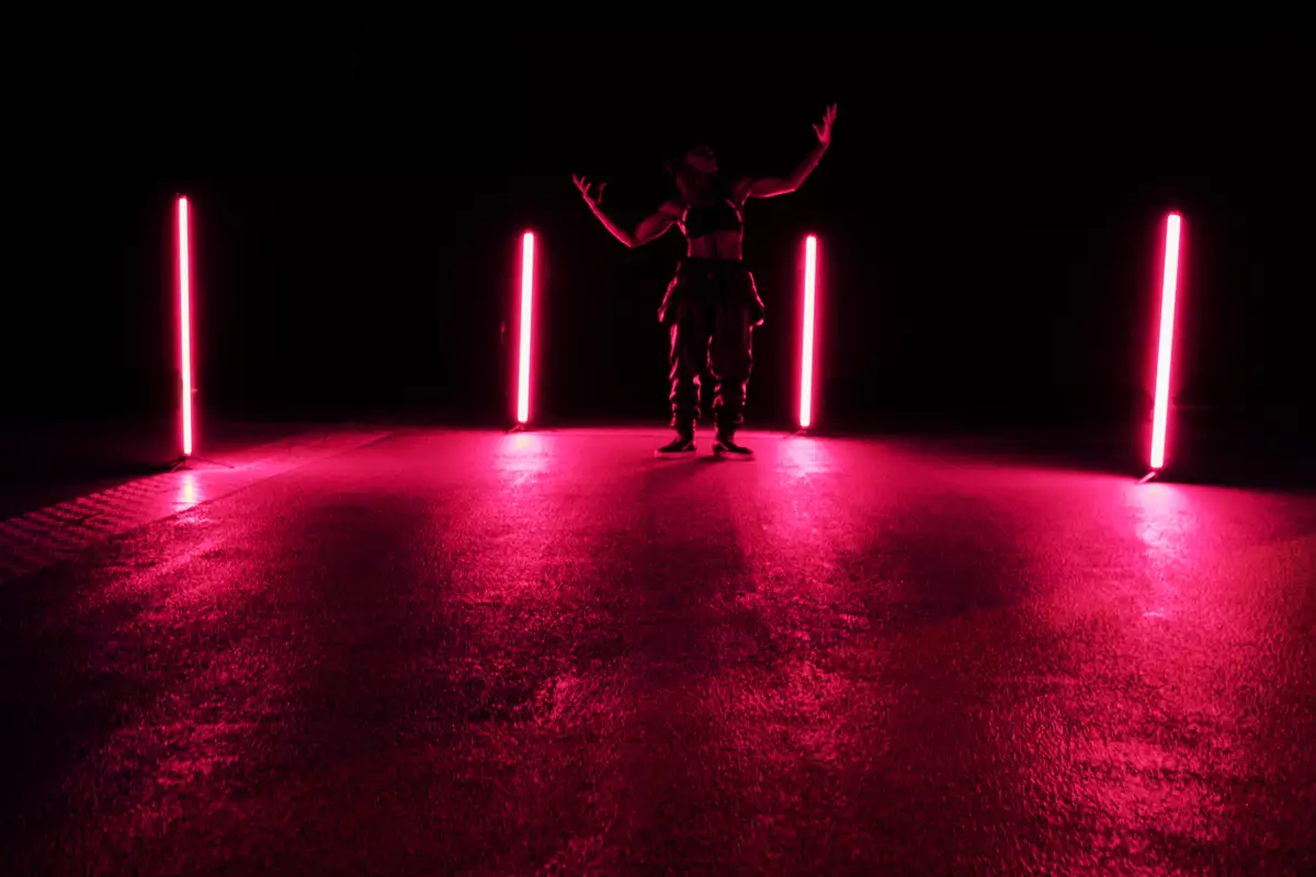 A person stands in a dimly lit space between four vertical neon lights emitting a pink glow, their arms raised in a dramatic pose