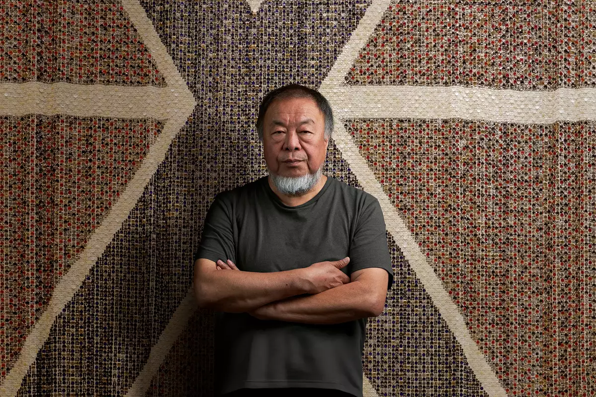 Ai Weiwei stood with arms folded wearing a black t-shirt, in front of a giant flag made of buttons