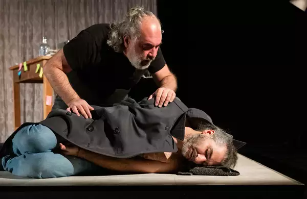 A person lying down on stage with eyes closed underneath a coat – with another person peering over him