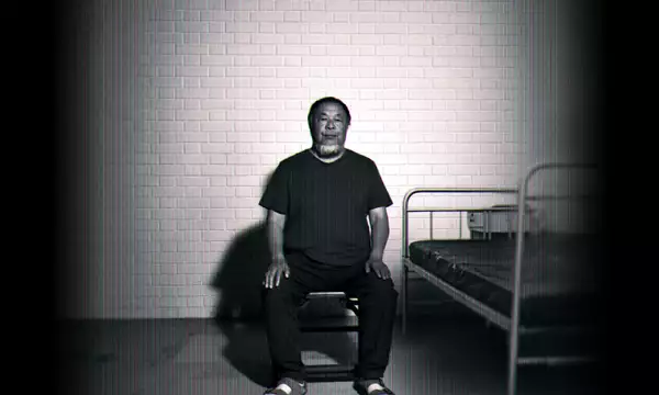 Black-and-white image of Ai Weiwei sitting on a chair beside a simple metal bed against a brick wall.
