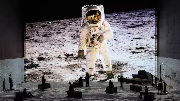 People sit and stand in a dark immersive exhibition space with a massive floor-to-ceiling projection of an astronaut walking on the moon. The lunar surface image extends across the floor and walls, creating a sense of being on the moon.