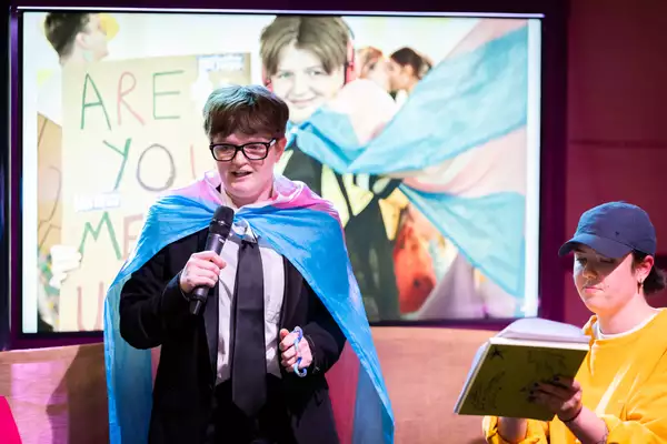 A performer at Balmy Army: A Contact Takeover wearing a cape and speaking into a microphone