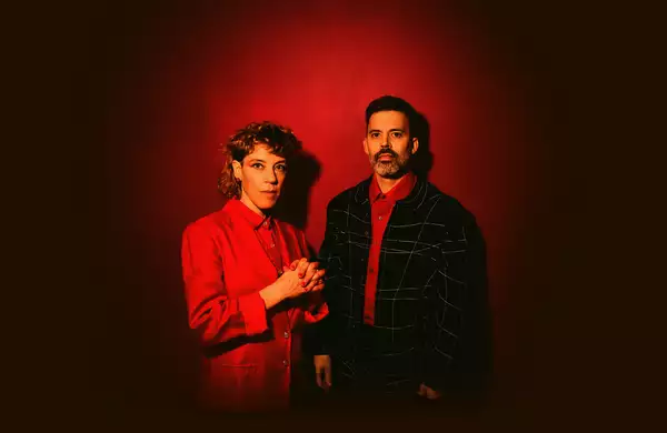 Two members of tune-yards wearing suits on a red background. The image features vibrant colours and dramatic red lighting.