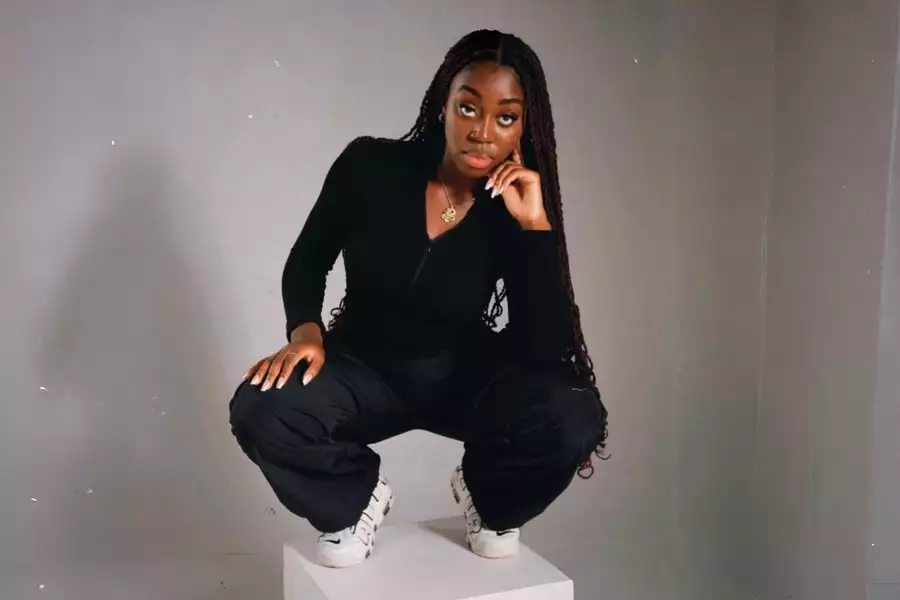 EKUA, dressed in black with white sneakers, poses thoughtfully on a white cube against a gray background.